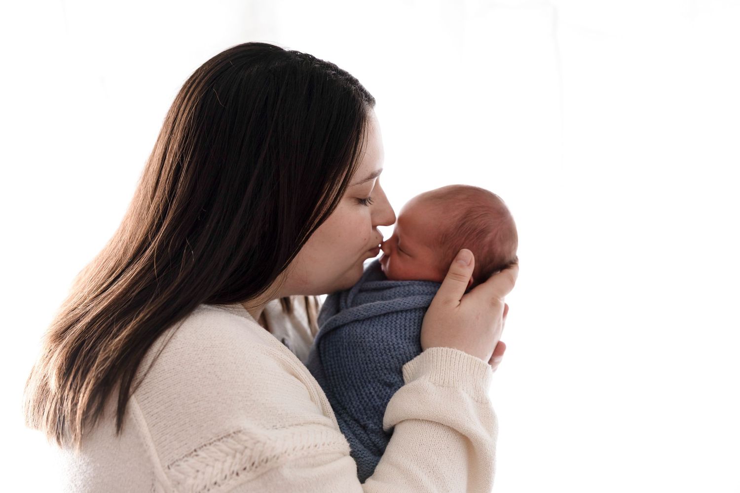 A mother tenderly kisses her newborn baby, cradling the infant gently against a soft white background.
