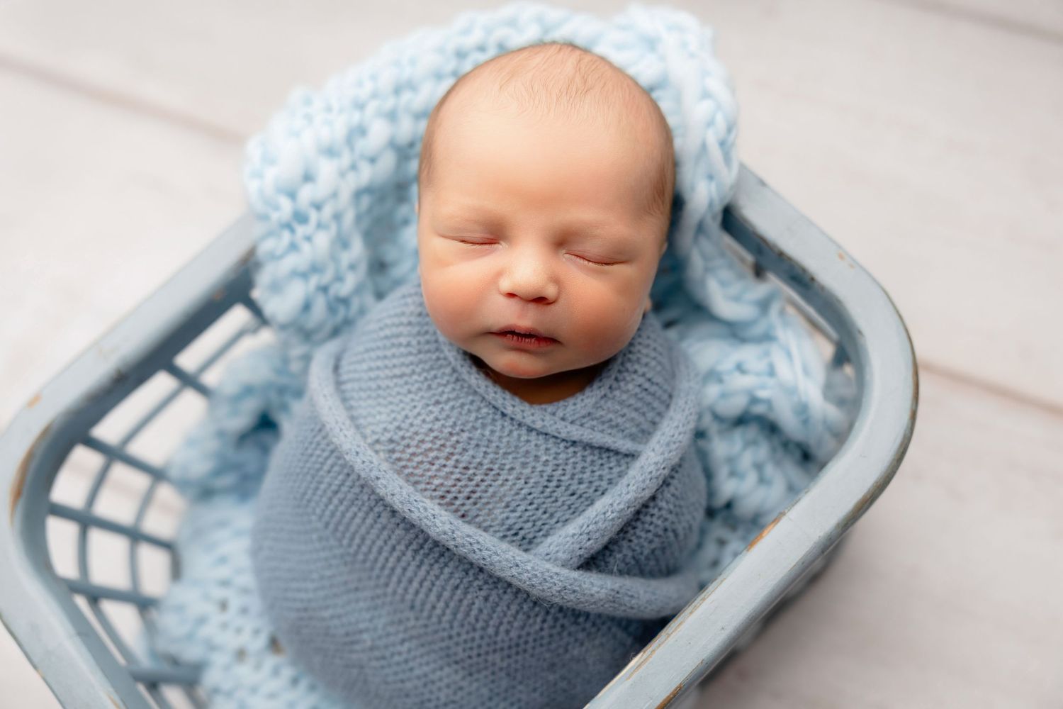 Newborn baby swaddled in gray knit wrap sleeping peacefully in a blue basket with soft blue blanket.