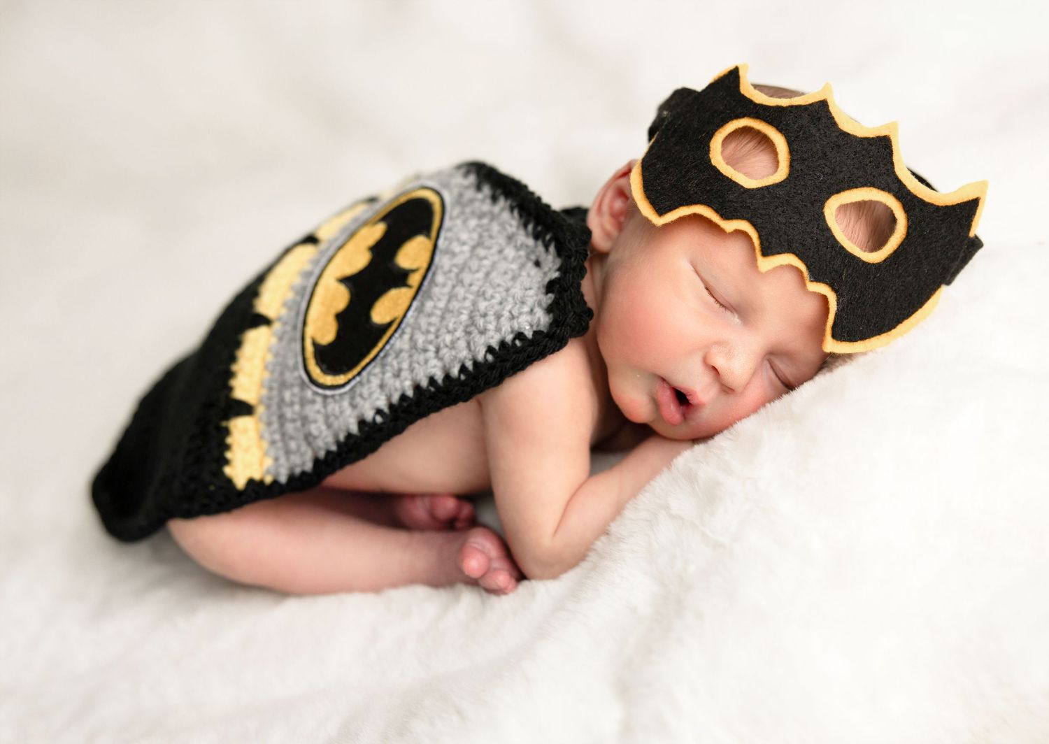 Sleeping newborn baby dressed in a crochet Batman costume with cape and mask hat on white blanket.