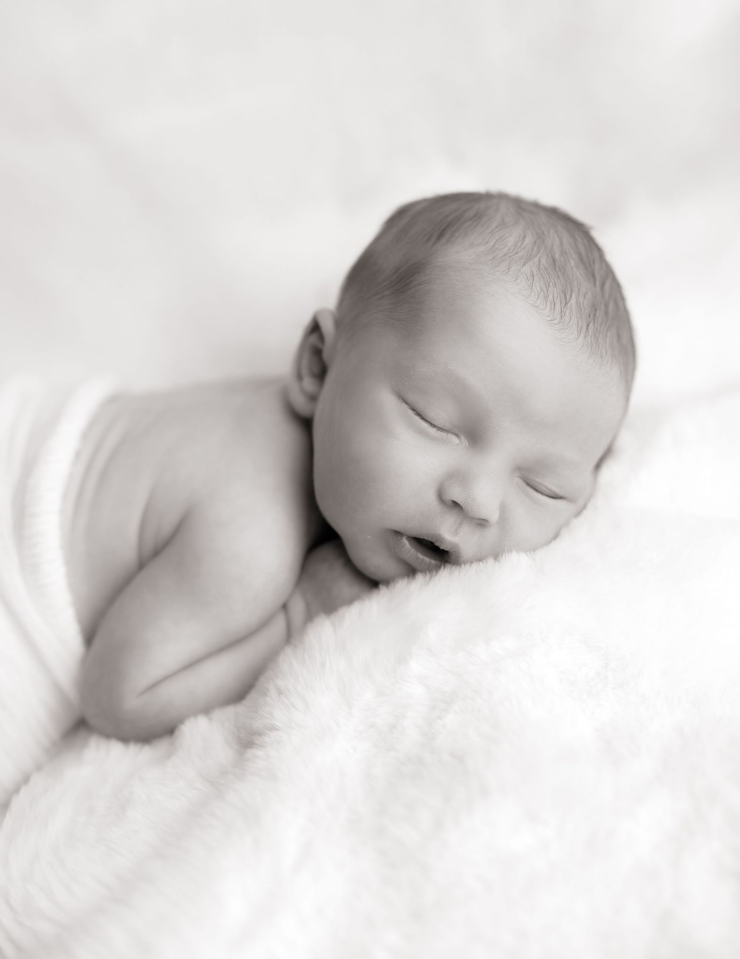 Black and white newborn baby sleeping peacefully on a soft white fluffy blanket in a serene portrait photo.