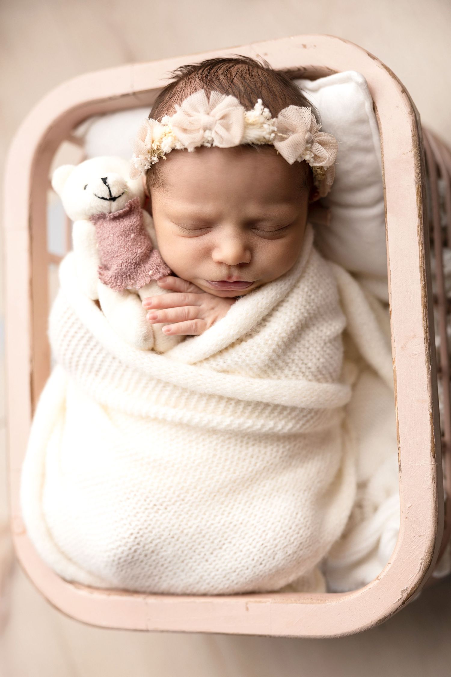 Sleeping newborn wrapped in white blanket with floral headband in pink basket.
