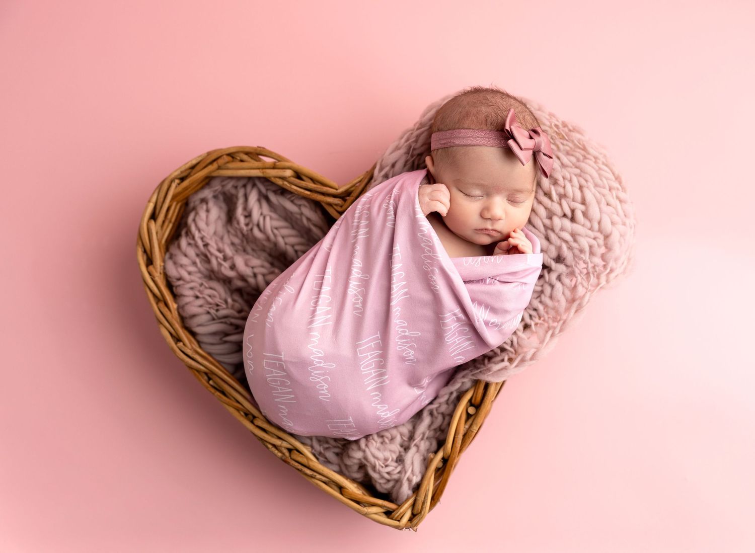 Sleeping newborn wrapped in pink inside heart-shaped basket.