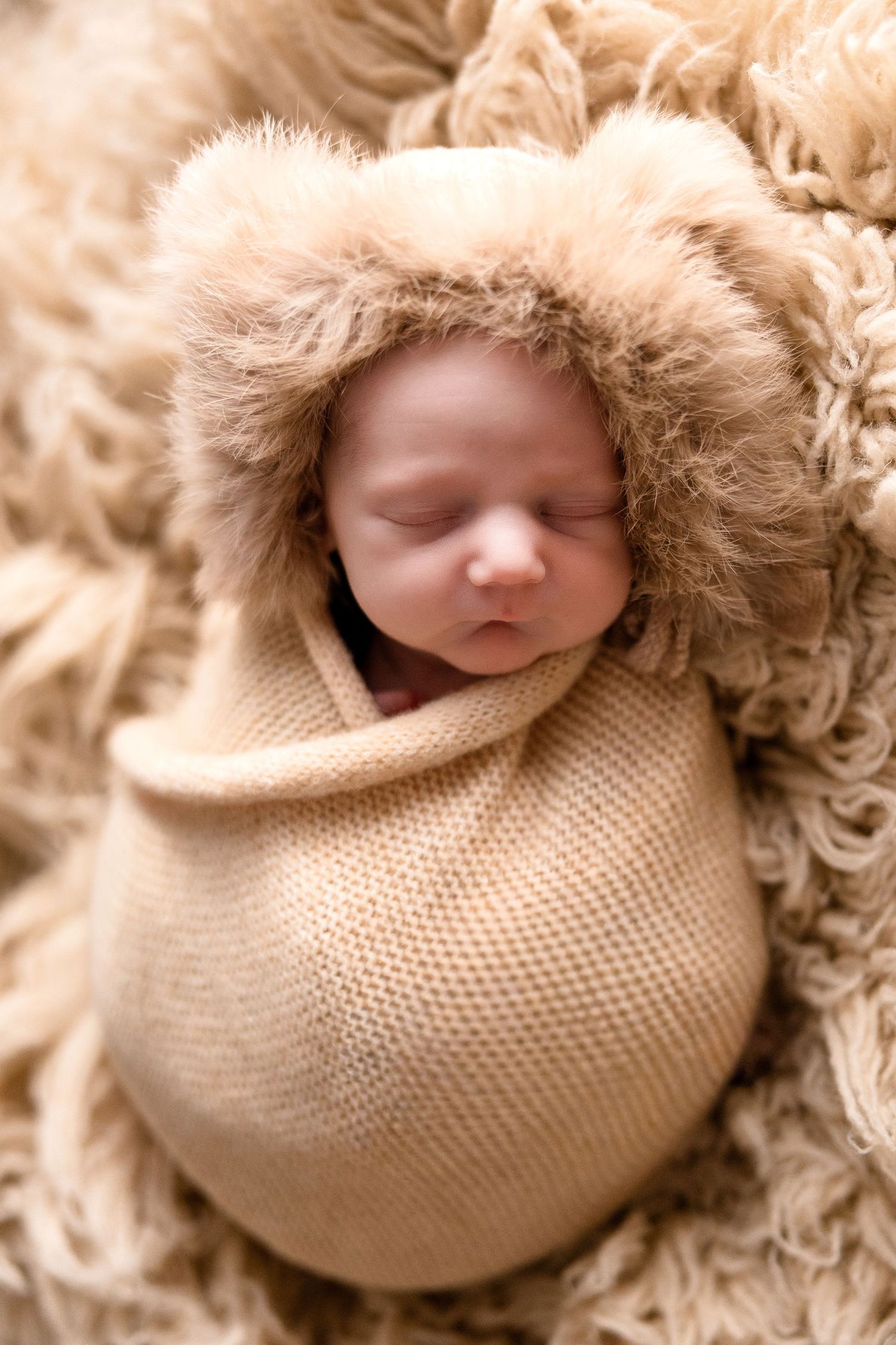 Sleeping newborn wrapped in cozy beige knit blanket with soft fur trim.