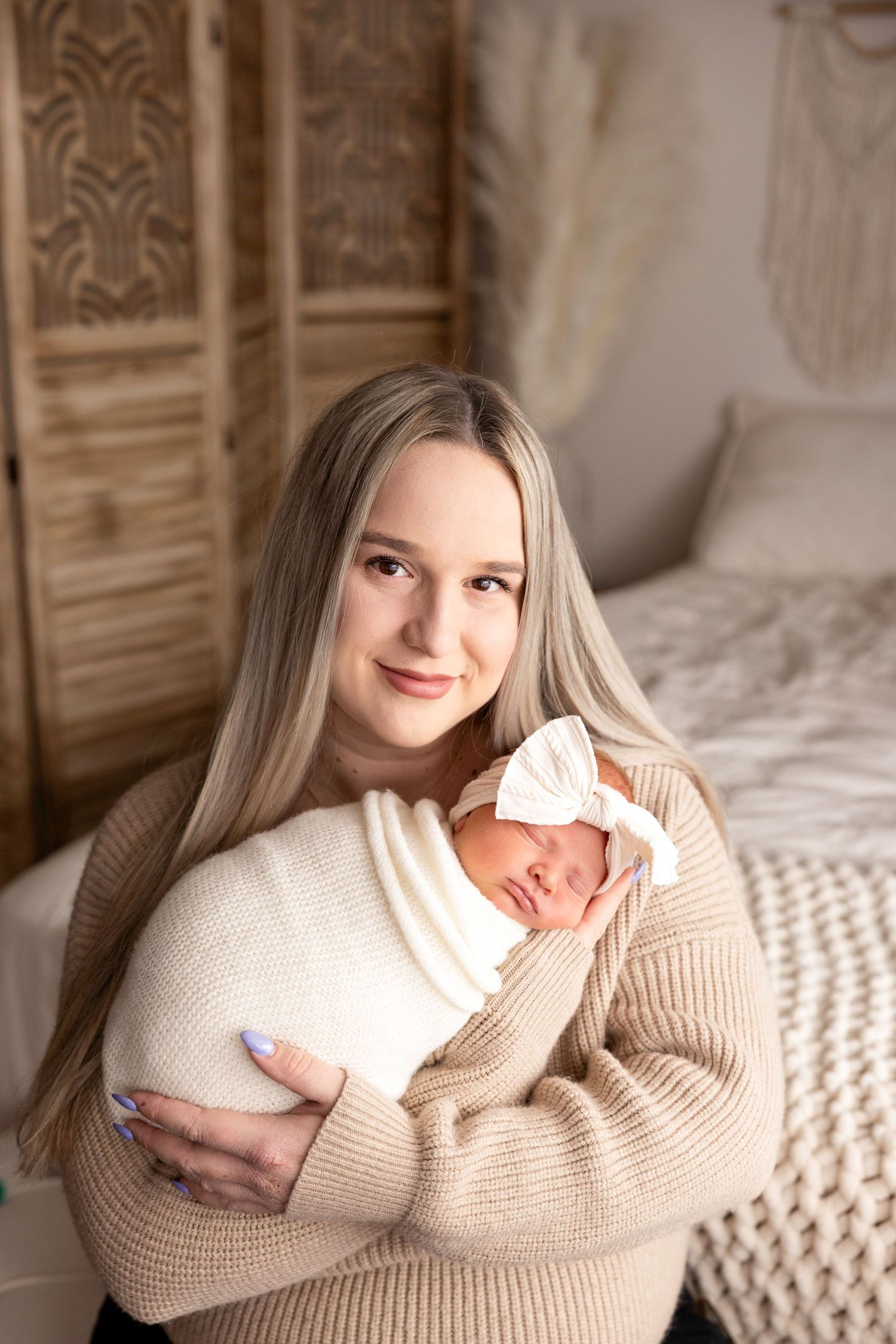 Smiling blonde mother in beige sweater cradling sleeping newborn wrapped in white swaddle blanket.