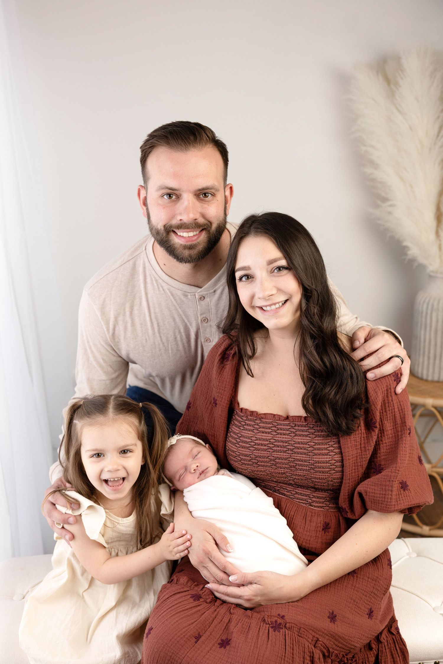 Happy family of four posing for a newborn portrait, parents smiling with toddler daughter and swaddled baby in a bright studio.