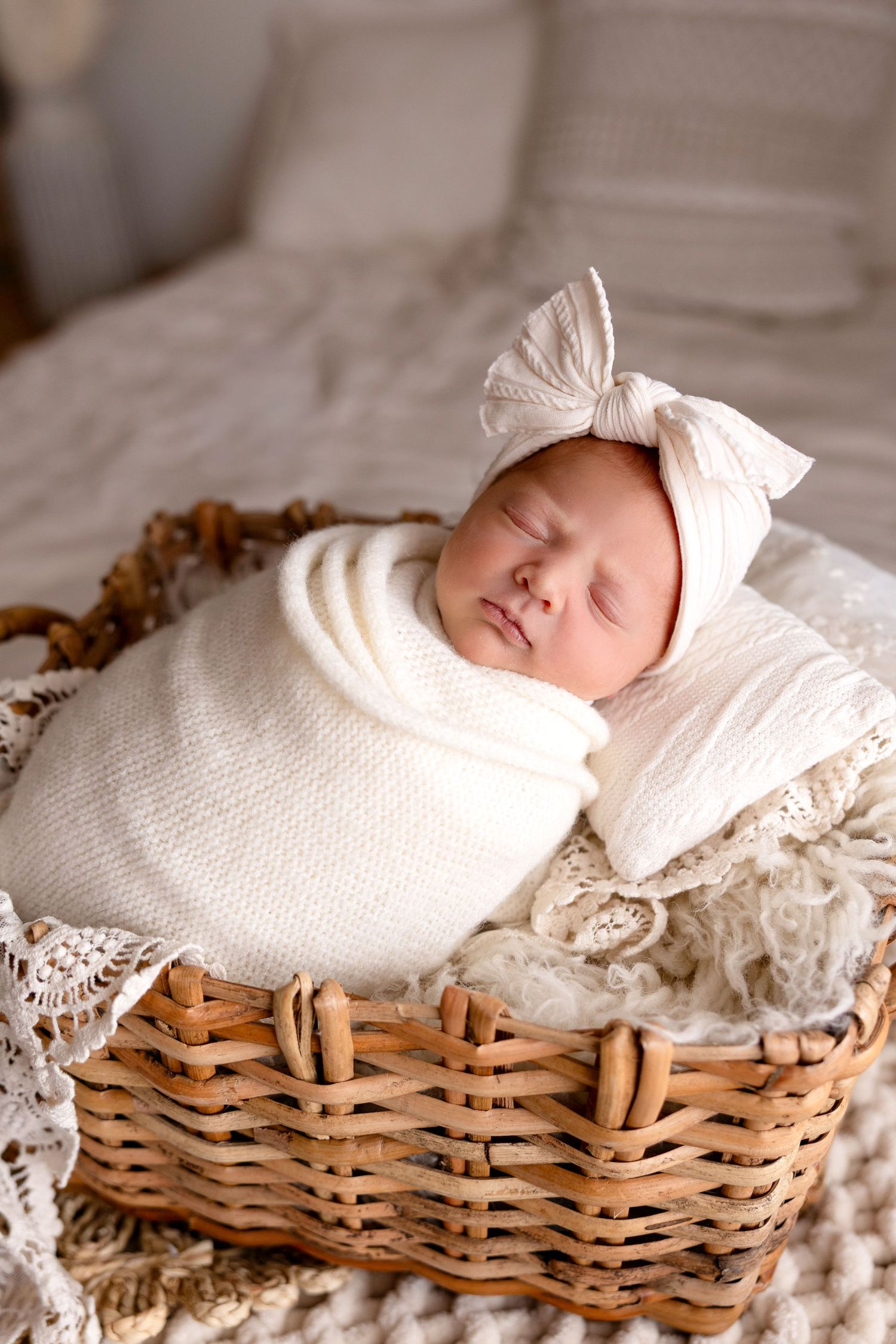 Sleeping newborn baby swaddled in white wrap wearing a bow headband, resting in a wicker basket.