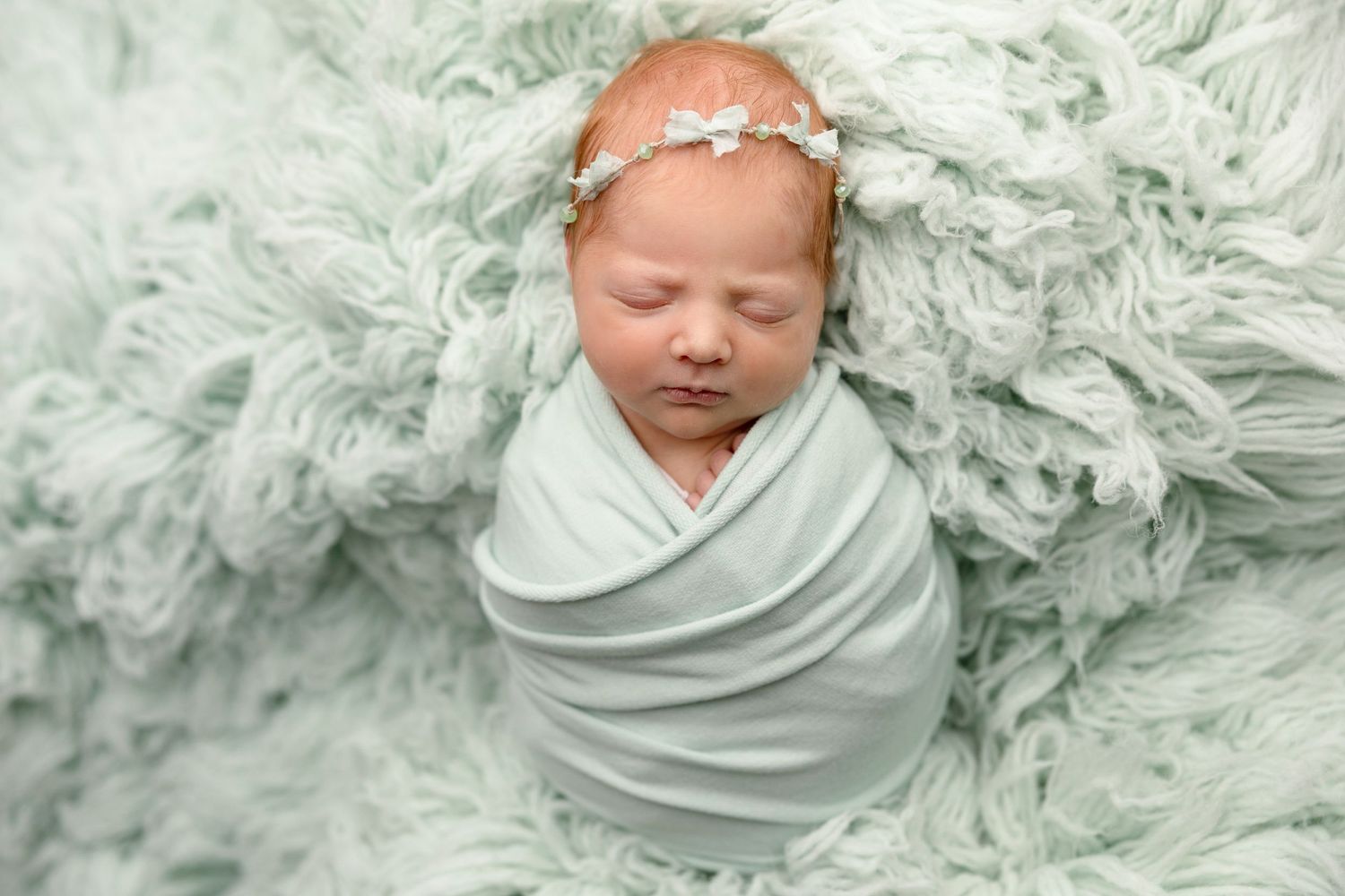 Newborn baby girl swaddled in mint green wrap sleeping on fluffy sage rug wearing delicate floral headband.
