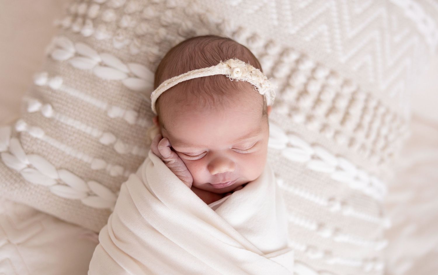 Peaceful newborn baby swaddled in white wrap, wearing a delicate headband, sleeping on a soft cream textured blanket.