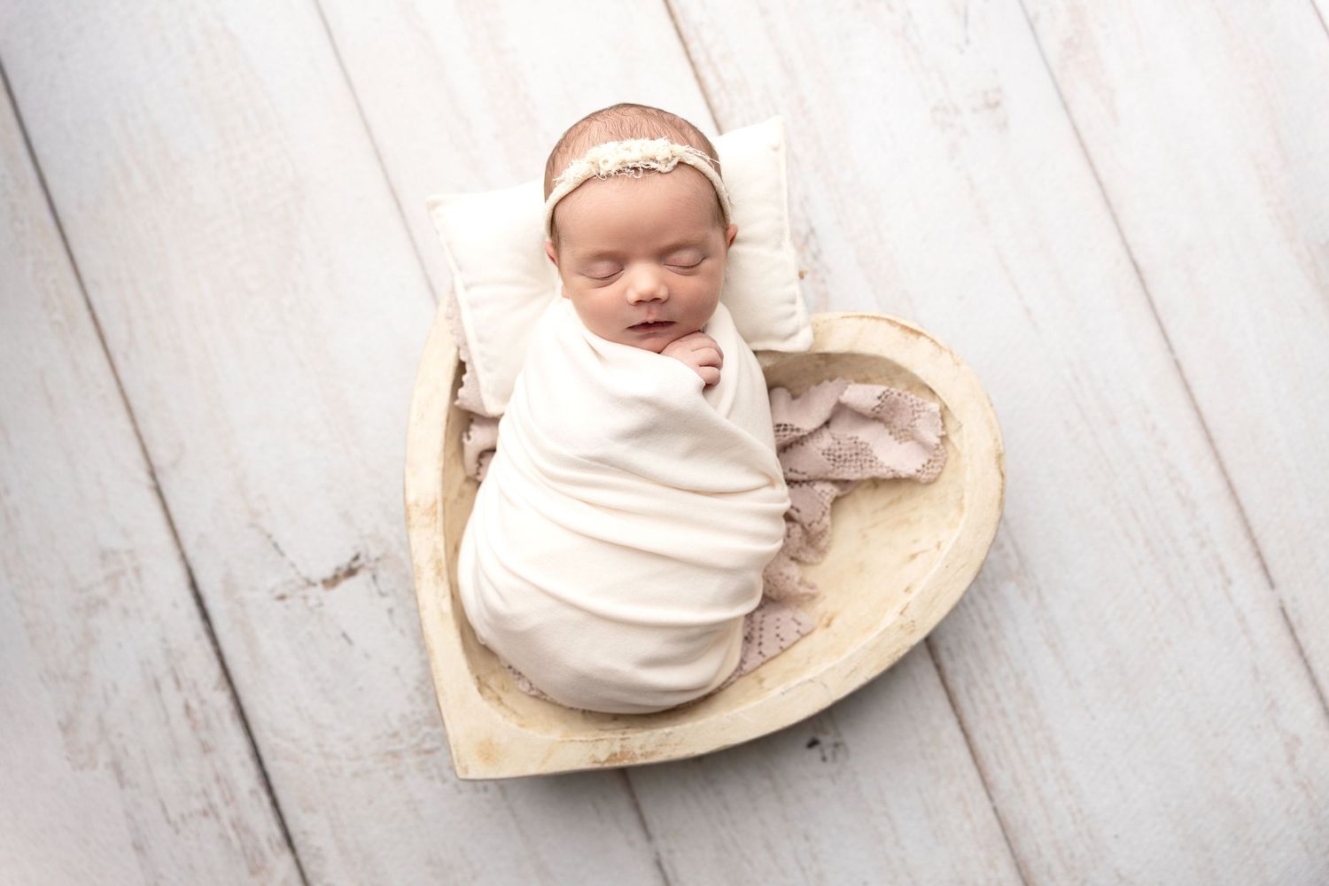 Newborn baby swaddled in white wrap sleeping in a wooden heart-shaped bowl with a floral headband.