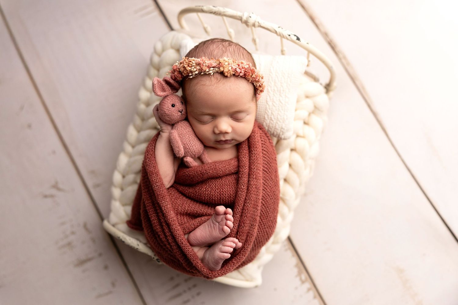 Newborn baby swaddled in rust wrap with floral headband sleeping peacefully in a white basket.