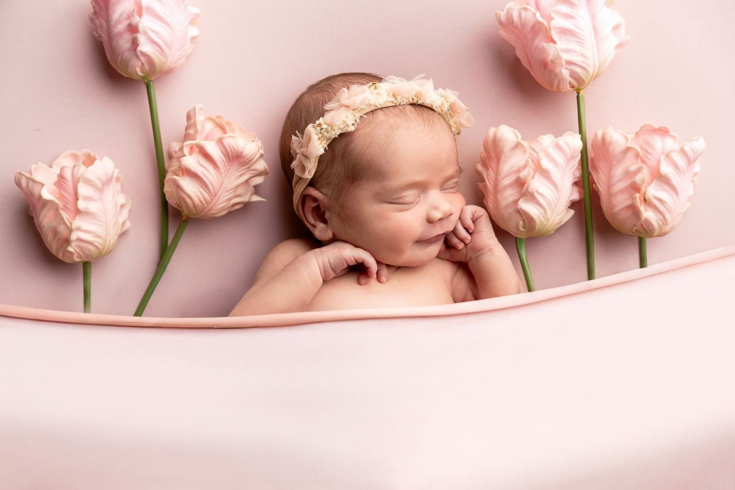 Sleeping newborn baby girl with floral headband surrounded by pink tulips on a soft pink background.