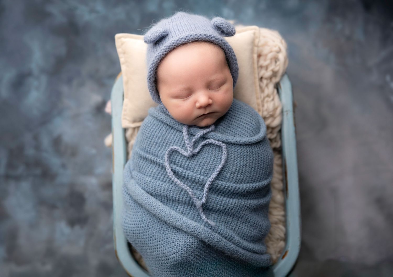 Sleeping newborn swaddled in soft blue wrap against dark background.