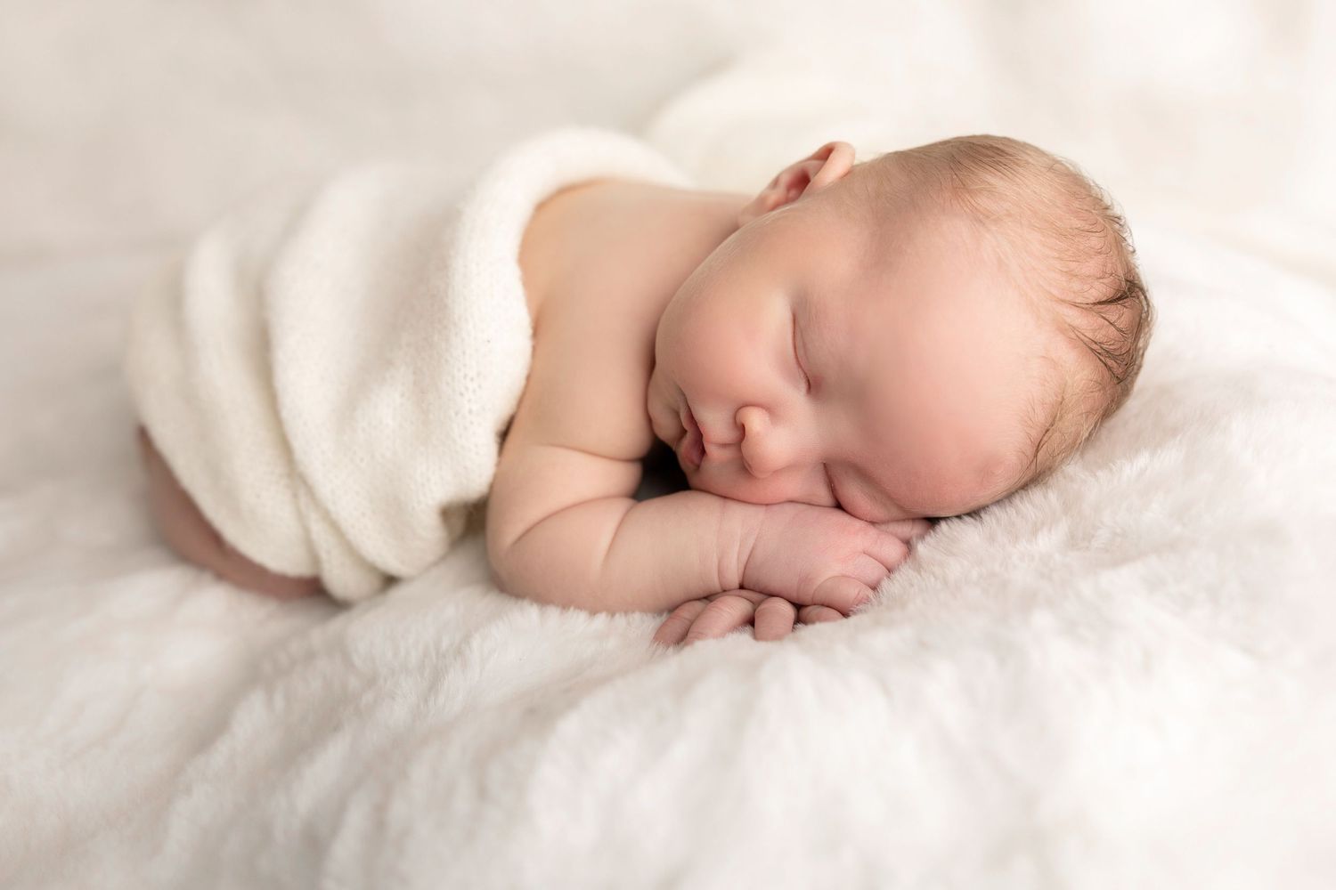 Sleeping newborn in white wrap on cream blanket.