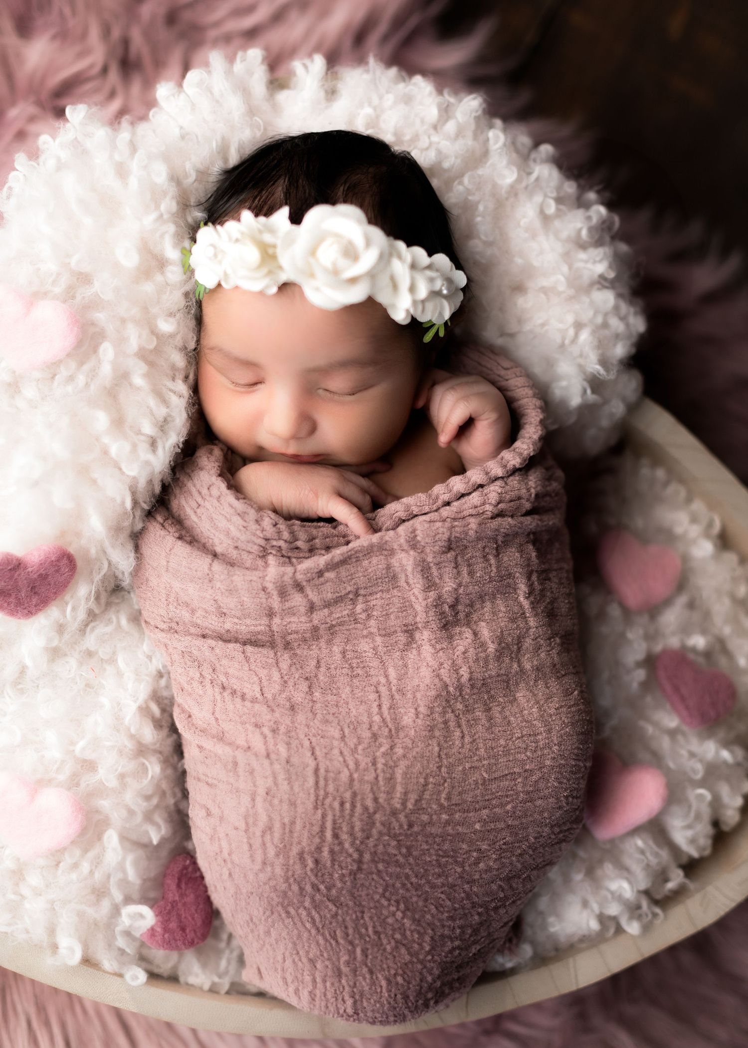 Sleeping newborn wrapped in pink swaddle with white floral headband on fluffy white blanket.