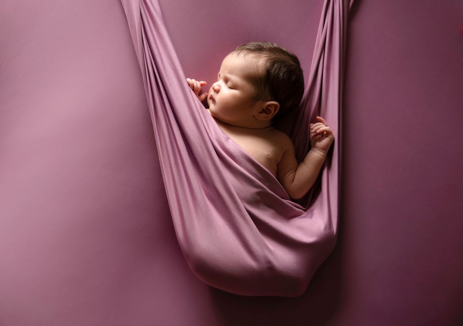 Artistic series showing sleeping baby suspended in pink fabric against burgundy backdrop.