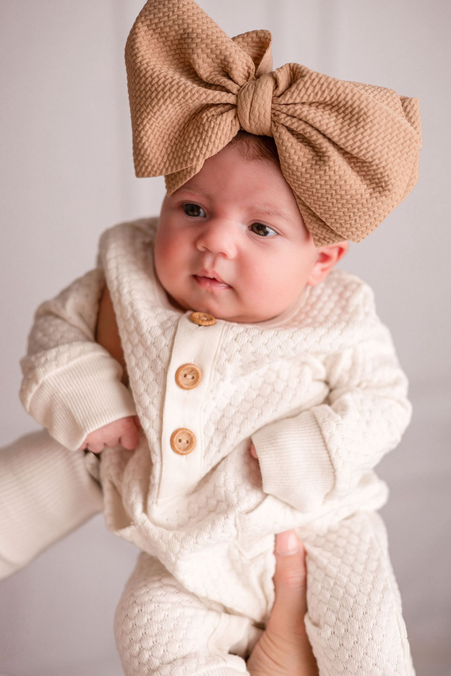 Baby wearing cream knit outfit with tan bow headband in studio portraits.