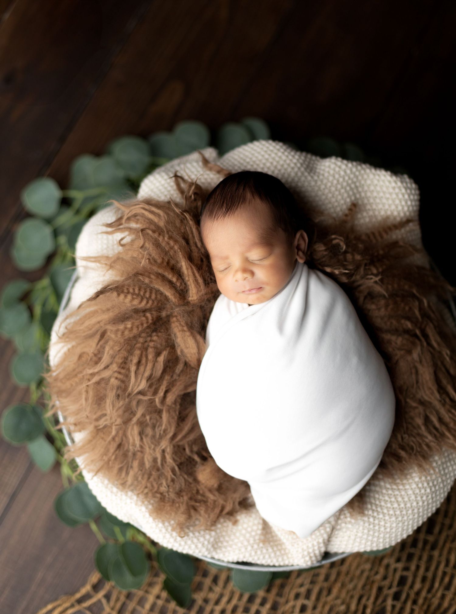 Newborn wrapped in white swaddle on brown fur with eucalyptus leaves.