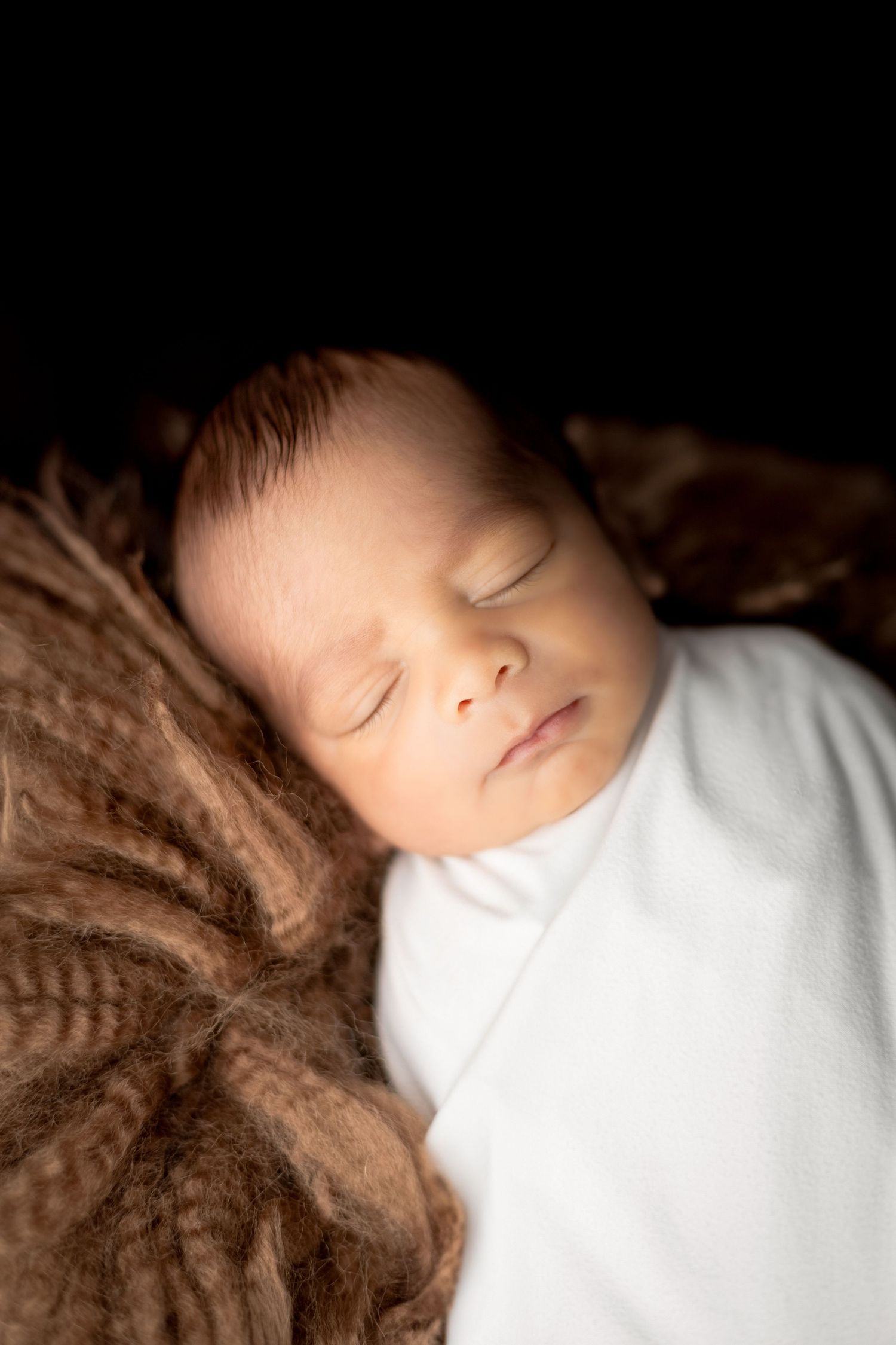 Peaceful sleeping newborn in white outfit on brown textured blanket.