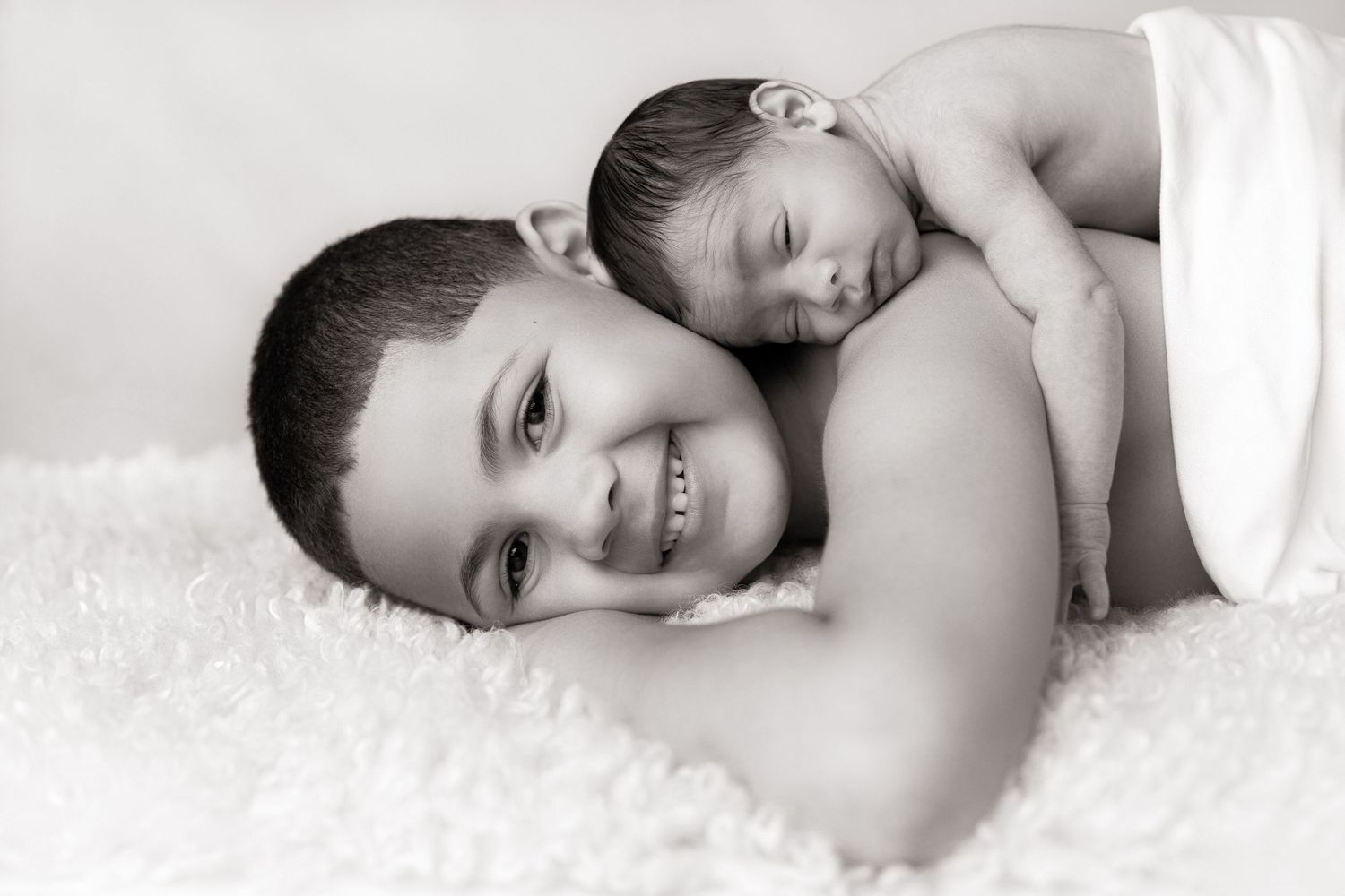 A tender black and white portrait of parent and sleeping newborn lying on soft white fur.