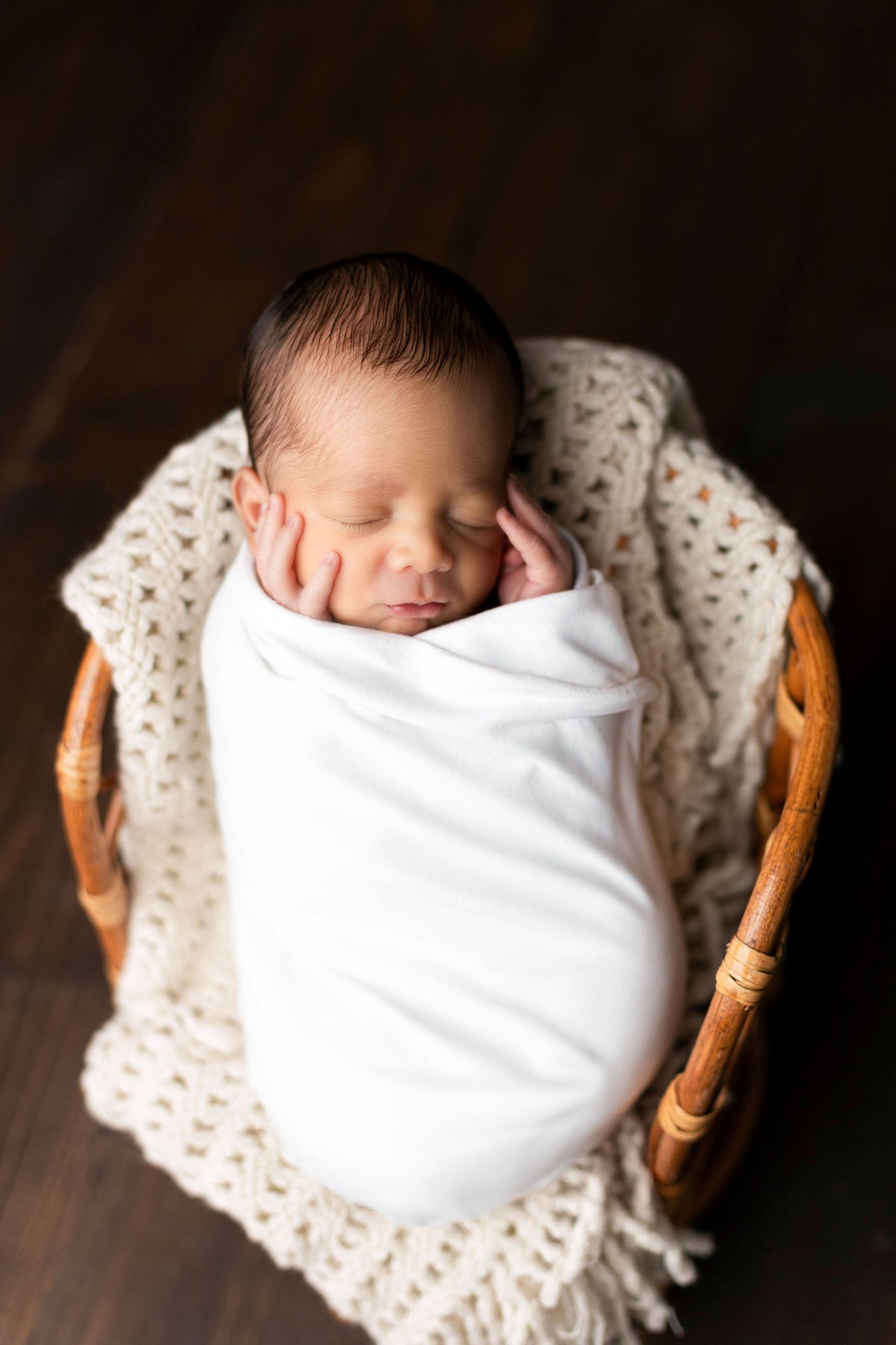 Sleeping newborn wrapped in white swaddle resting in wooden basket with knit blanket.