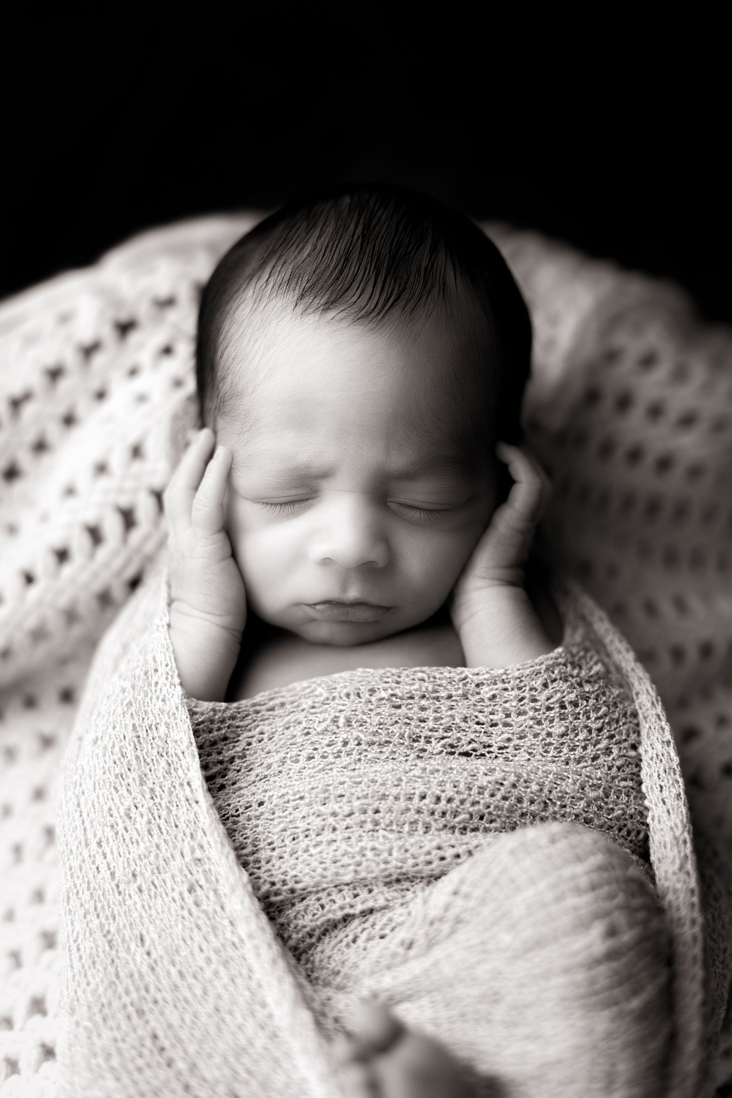 Artistic black and white portrait of sleeping newborn wrapped in knit blanket.