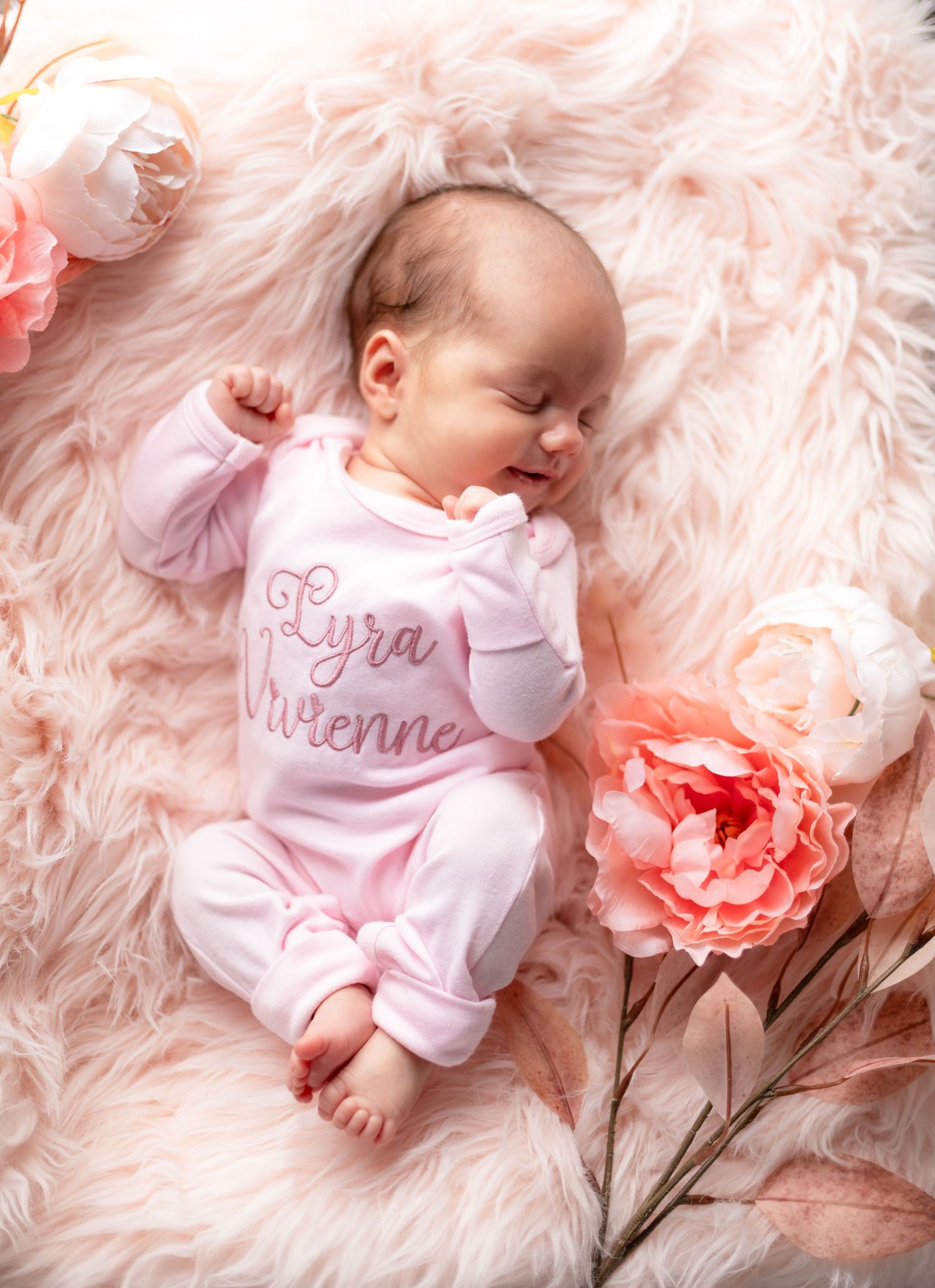 Sleeping baby in pink outfit on fluffy pink blanket with coral flowers.