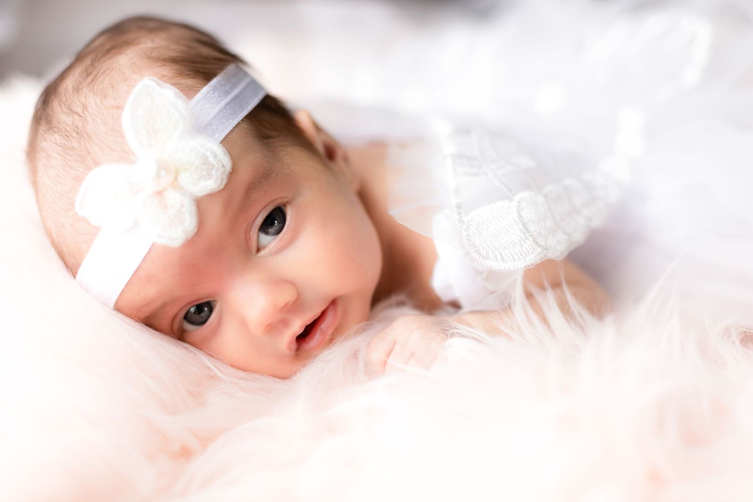 Baby in white lace outfit with bow headband on soft pink background.