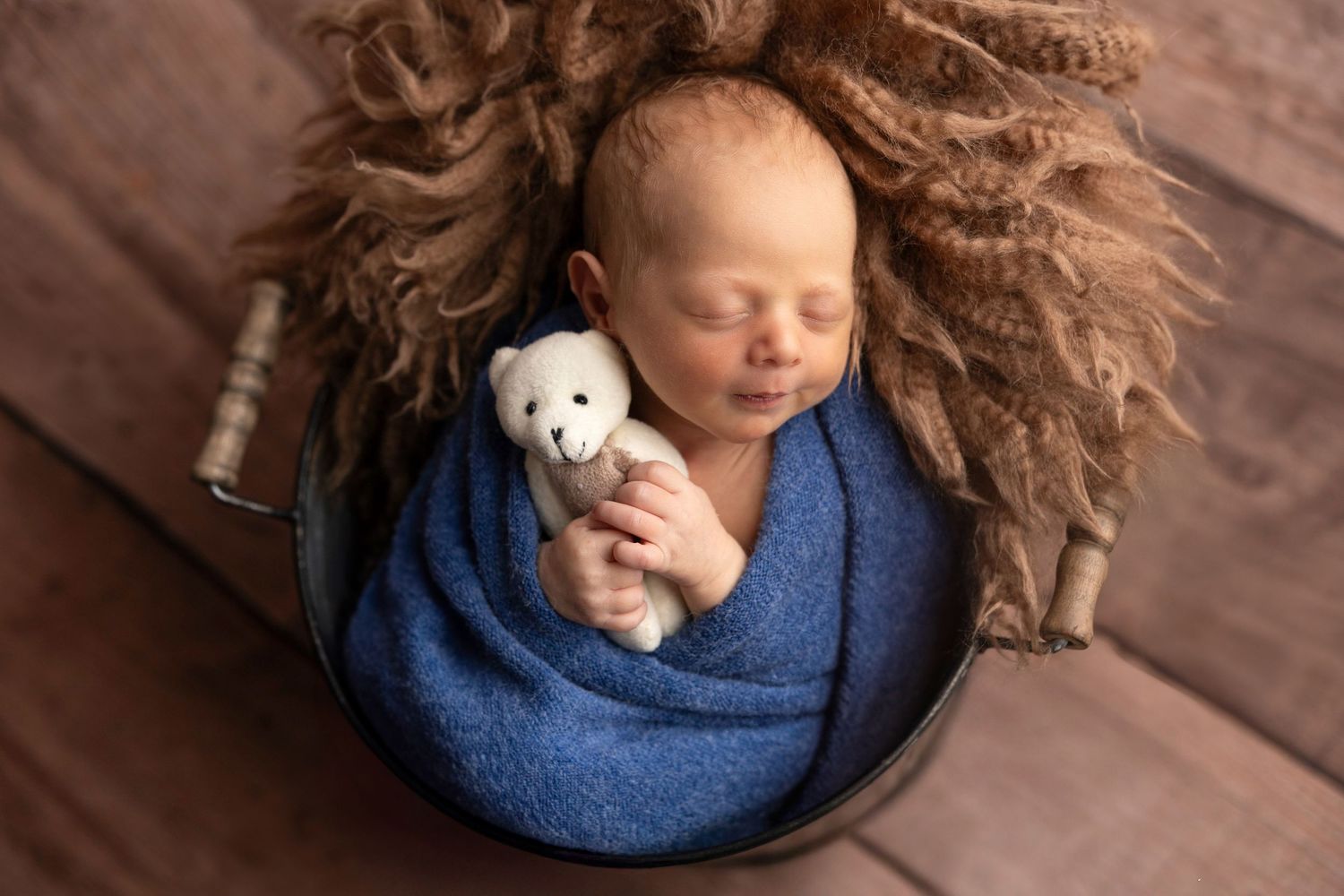 Baby resting on wooden floor with blue wrap and small white teddy bear.