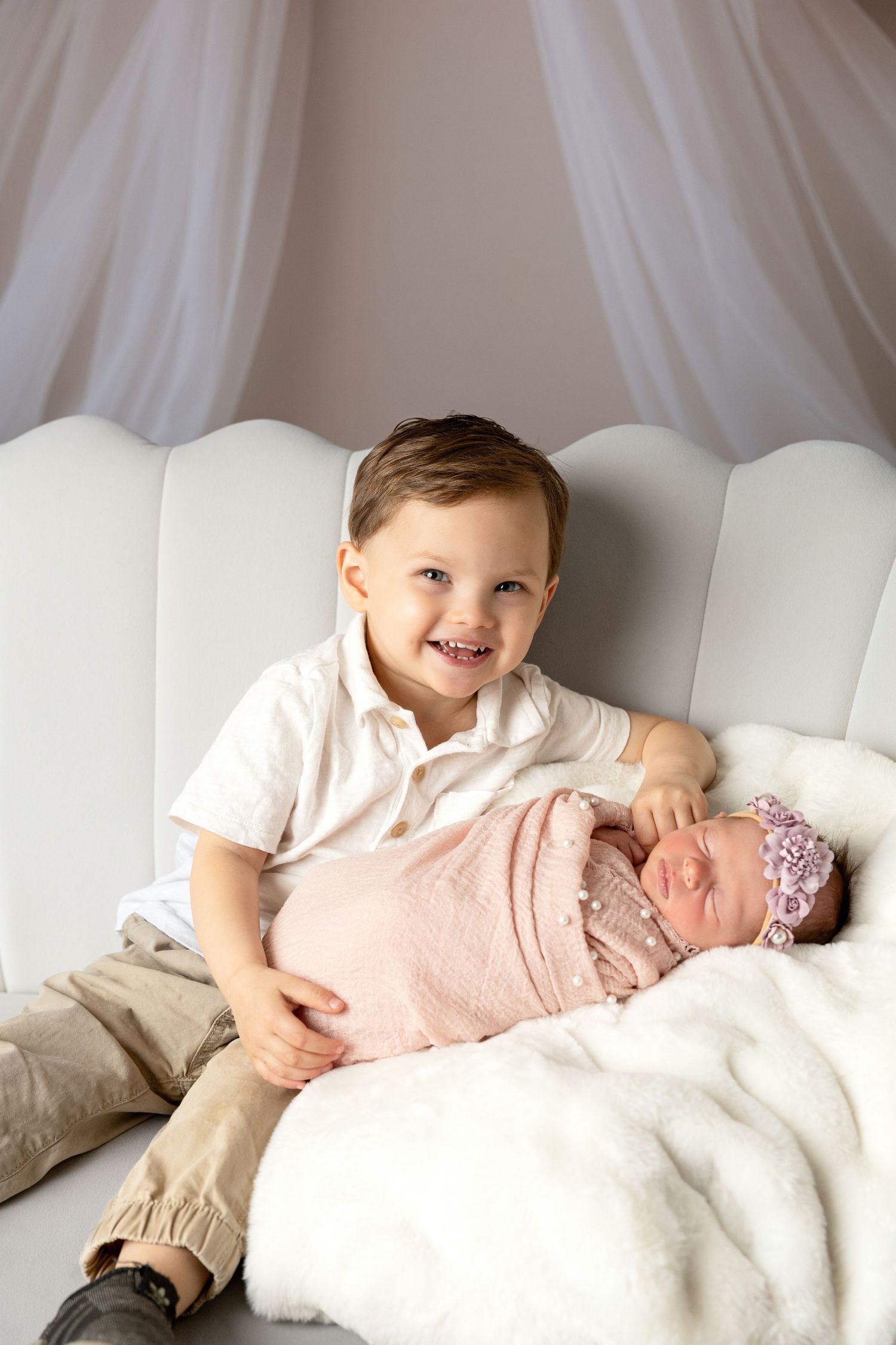 Sweet sibling photo session on white bed with soft pink accents.