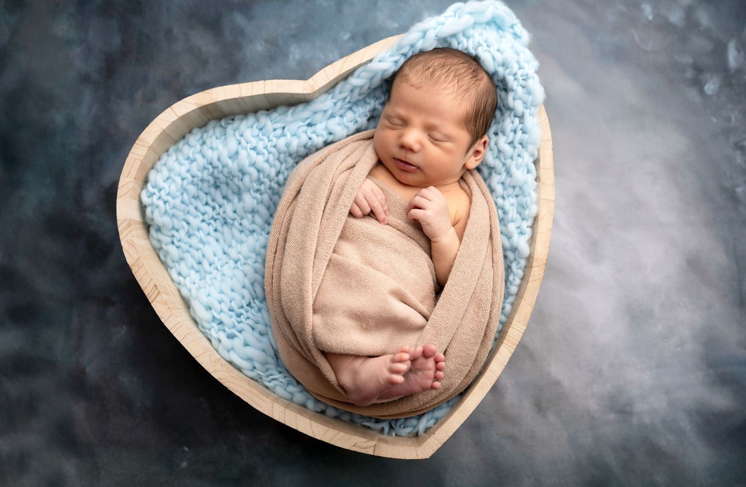 Sleeping newborn wrapped in beige fabric nestled in heart-shaped basket with blue blanket.