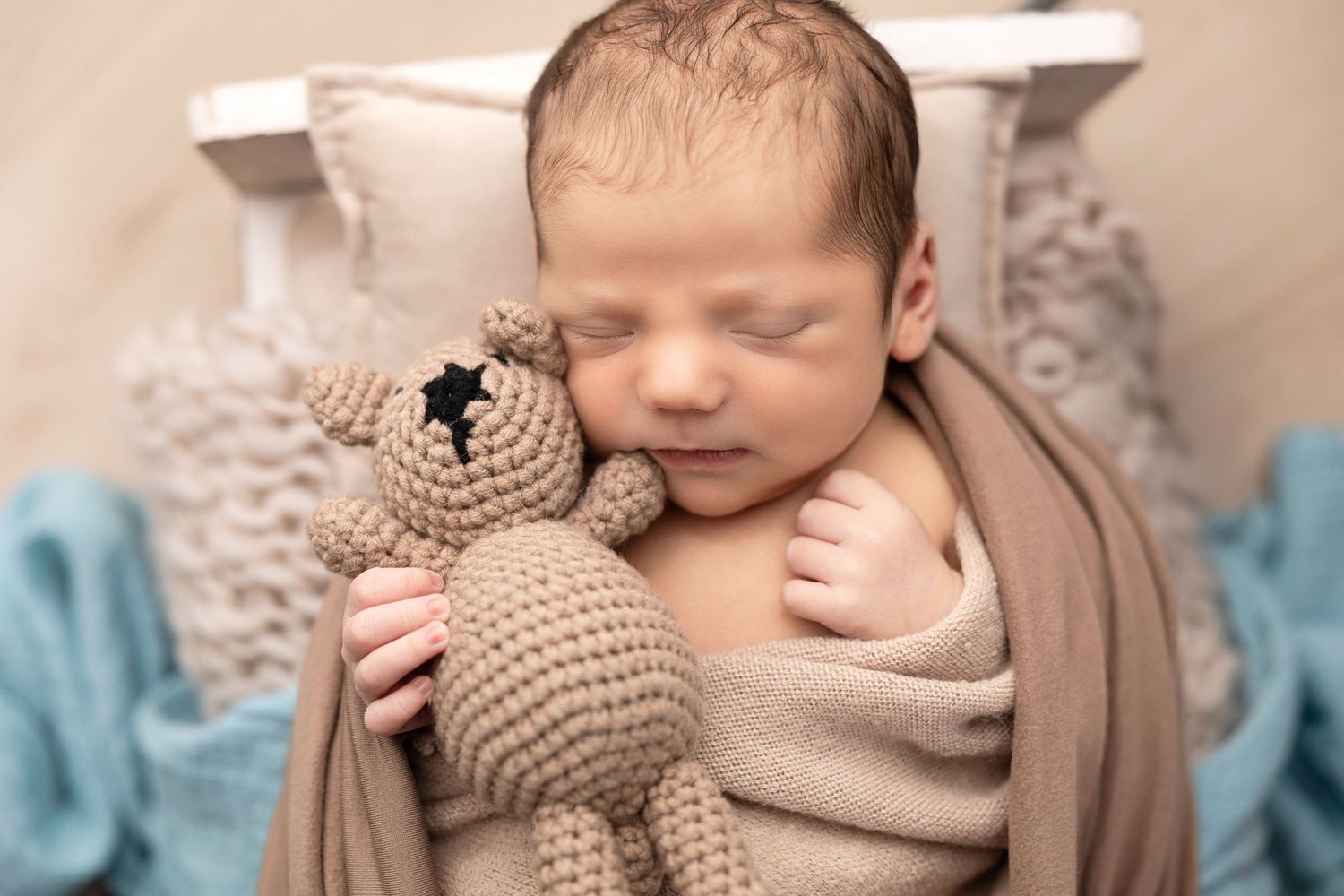 Sleeping newborn cuddling with knitted teddy bear wrapped in neutral toned swaddle.