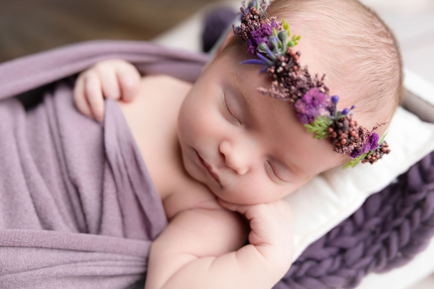 Sleeping newborn in purple outfit with floral crown during studio session.