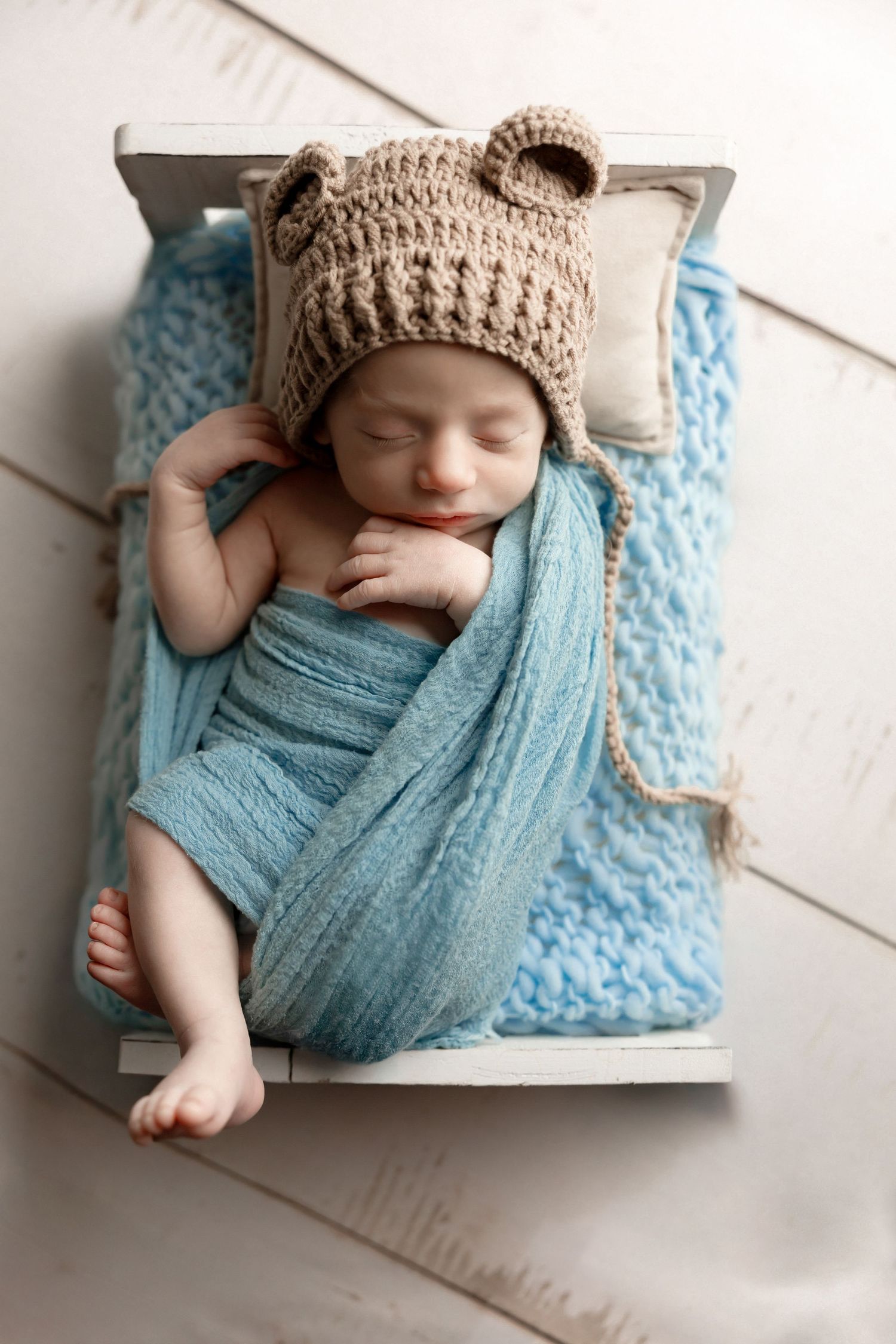 Sleeping baby wrapped in turquoise blanket wearing brown knit hat on textured surface.