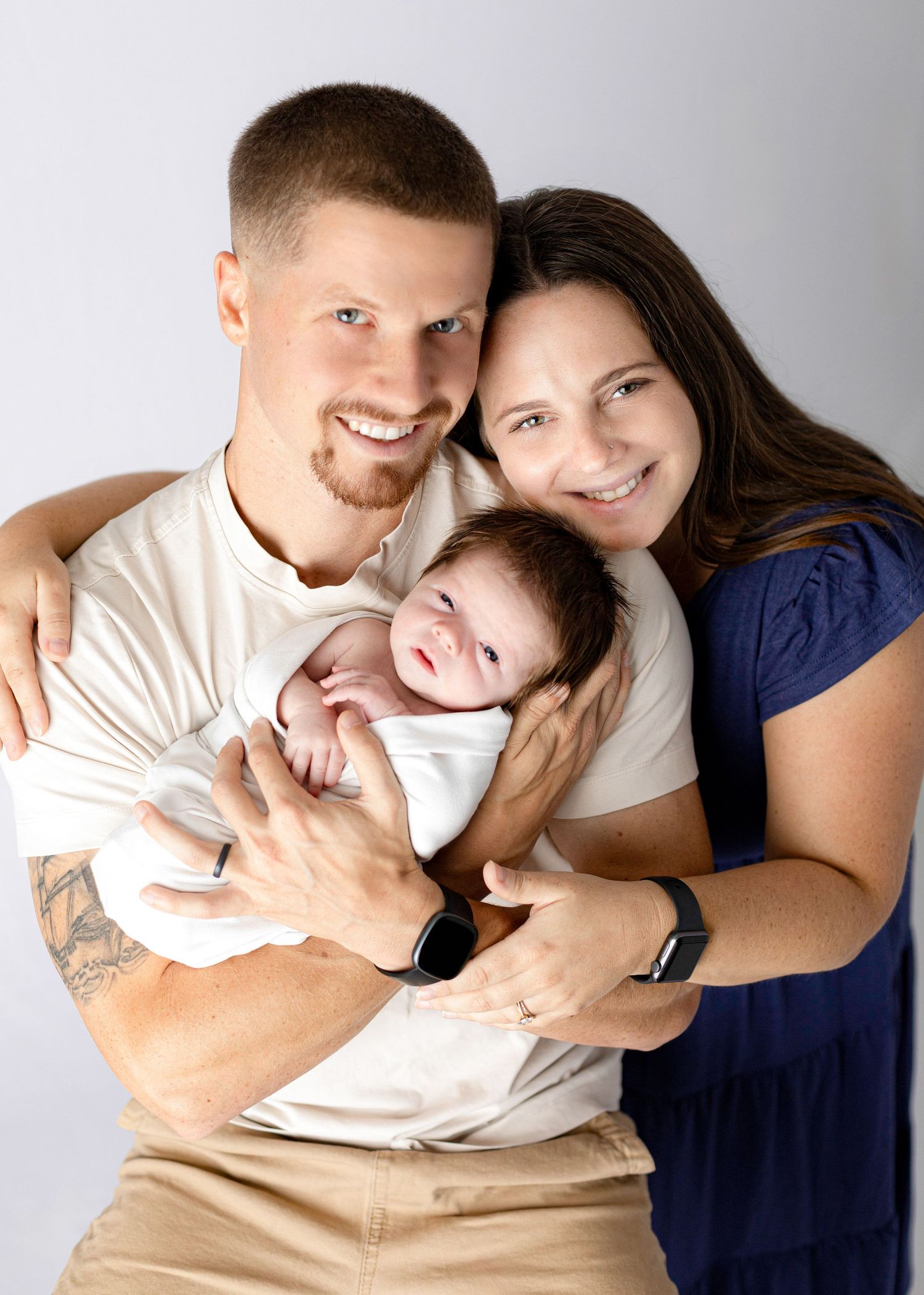 A loving family portrait with parents holding their newborn baby against a light background.