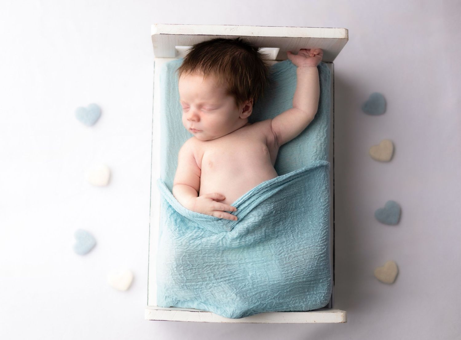 Sleeping newborn baby in light blue blanket lying in small white crib with heart decorations.