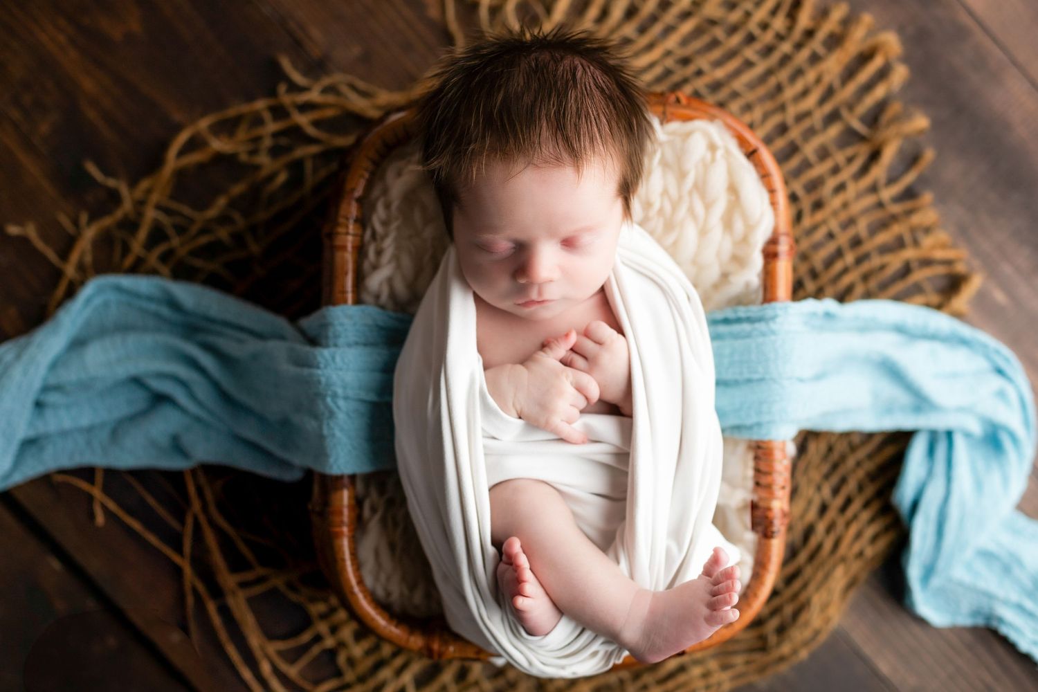Newborn baby sleeping in rustic wooden basket with blue and cream textiles.