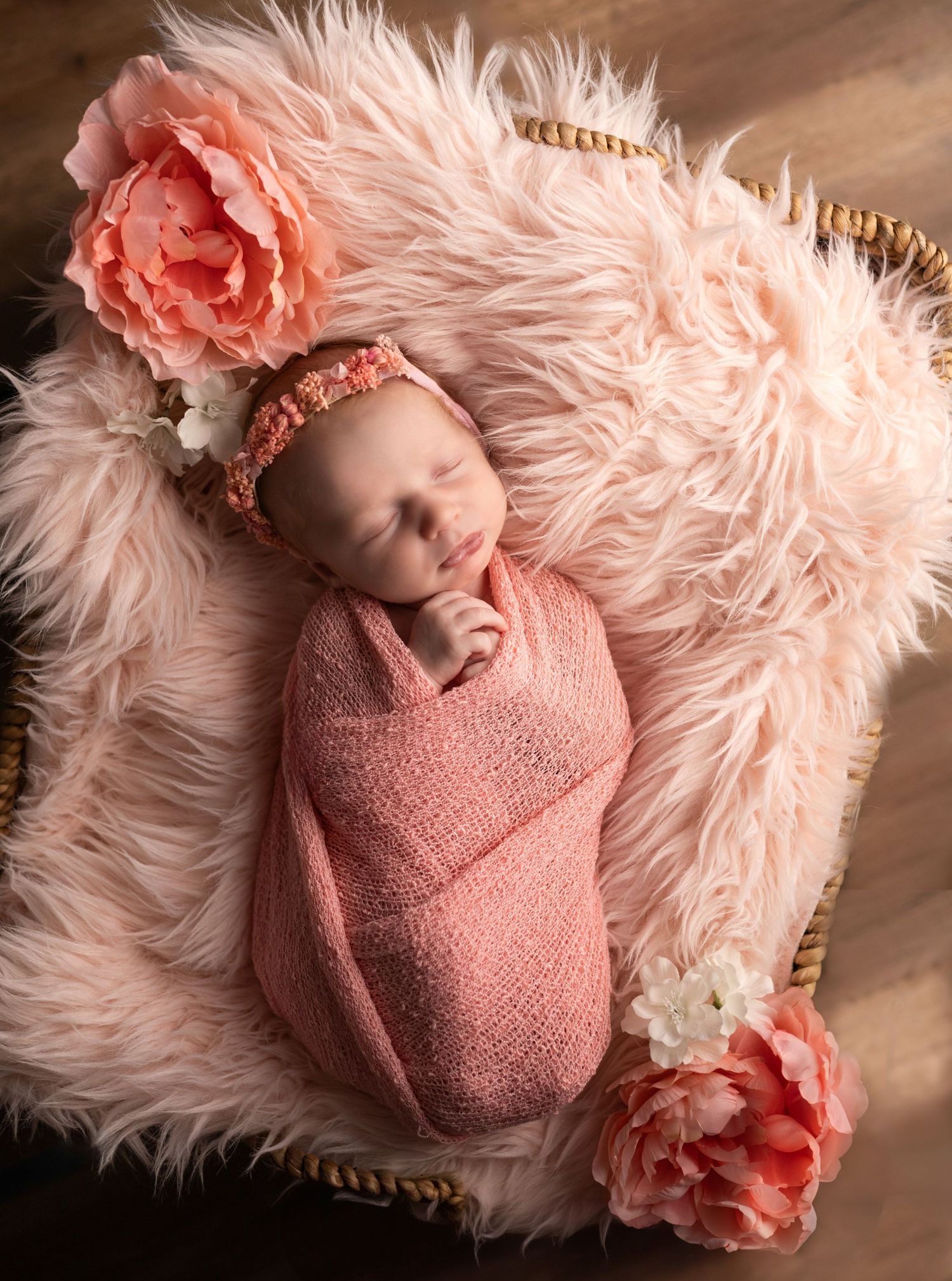 Newborn wrapped in pink swaddle on fluffy pink rug with coral flowers.