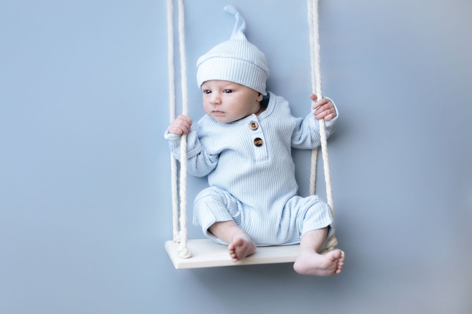 Baby in light blue outfit sitting on white wooden swing during photoshoot.