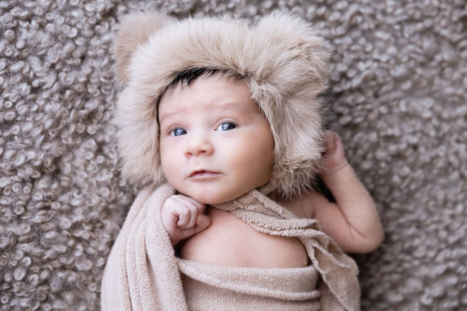 Newborn baby wearing a cozy fur hat lies on textured grey fabric.