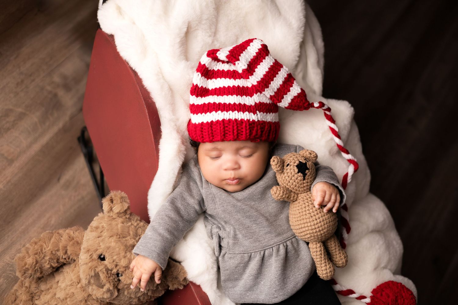 Baby in red and white striped hat rests with plush teddy bears on red chair.