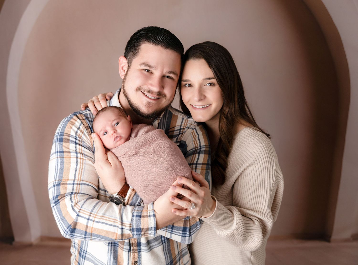 Intimate family portrait with a newborn baby wrapped in pink against a warm brown background.