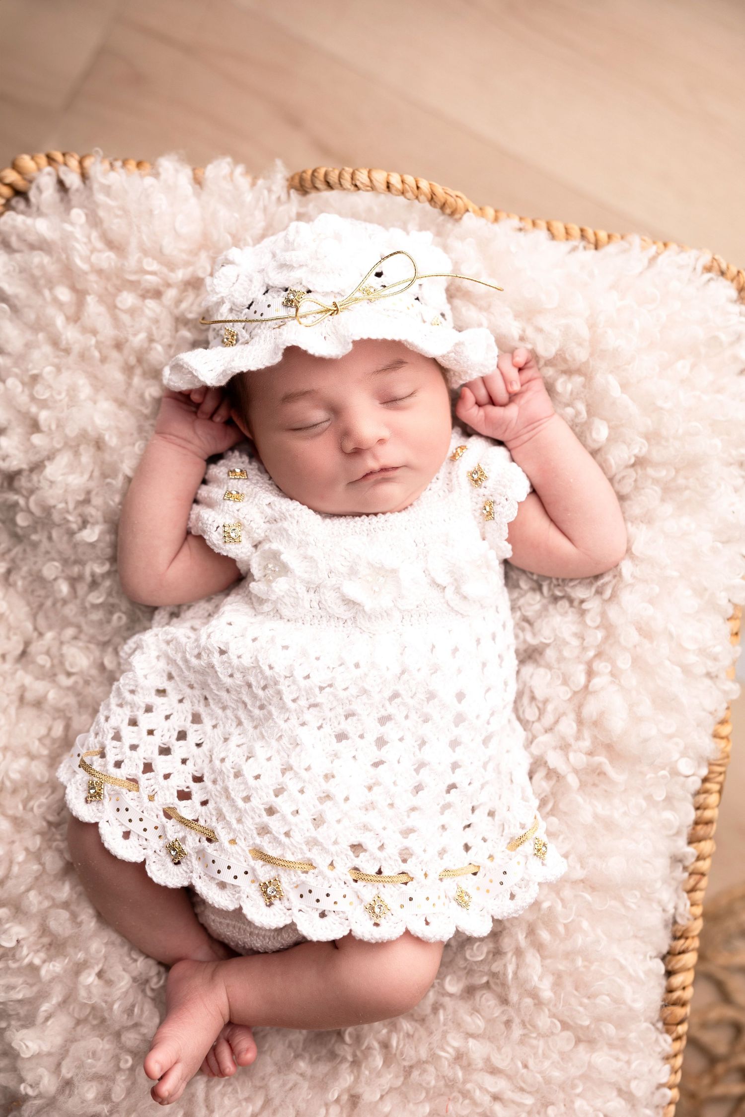 Sleeping newborn in white lace dress and bonnet on fluffy cream blanket.
