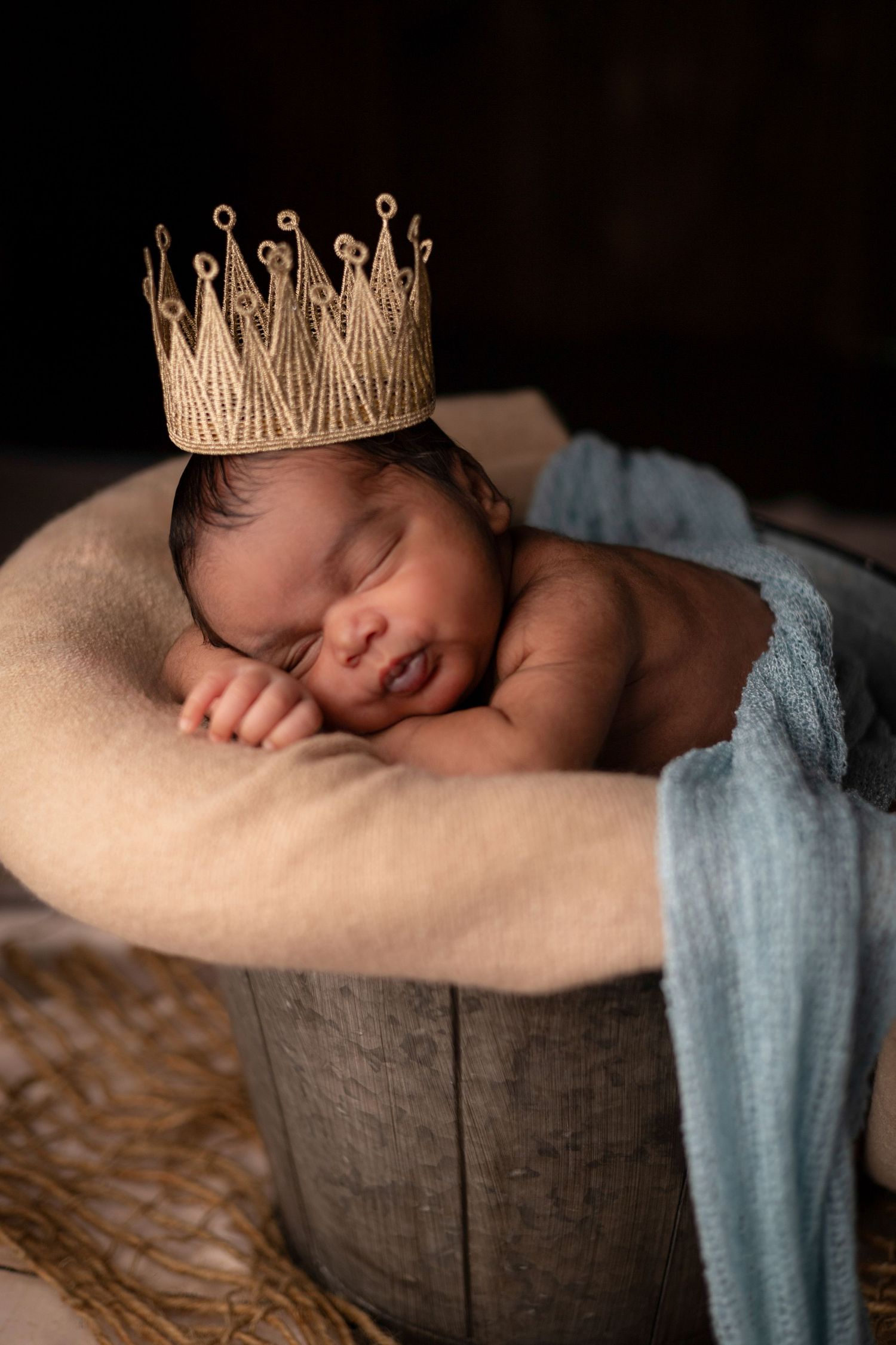 Sleeping newborn wearing gold crown rests on brown backdrop with blue fabric.