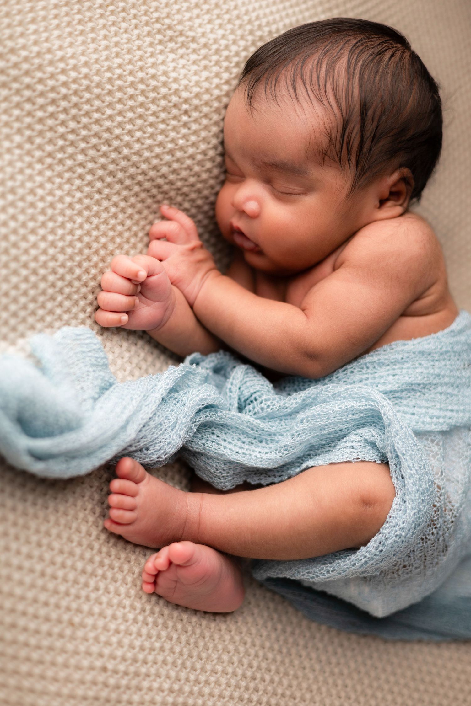 Newborn baby wrapped in light blue knit blanket sleeping on textured beige surface.