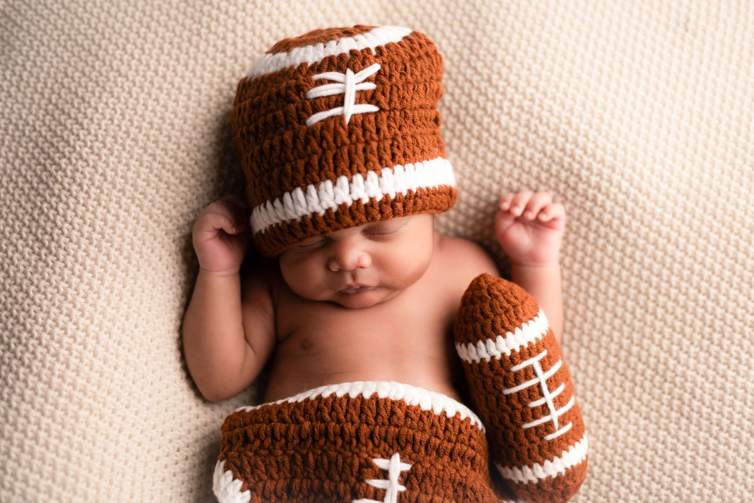 Newborn baby wearing brown football-themed knit outfit sleeping on cream background.