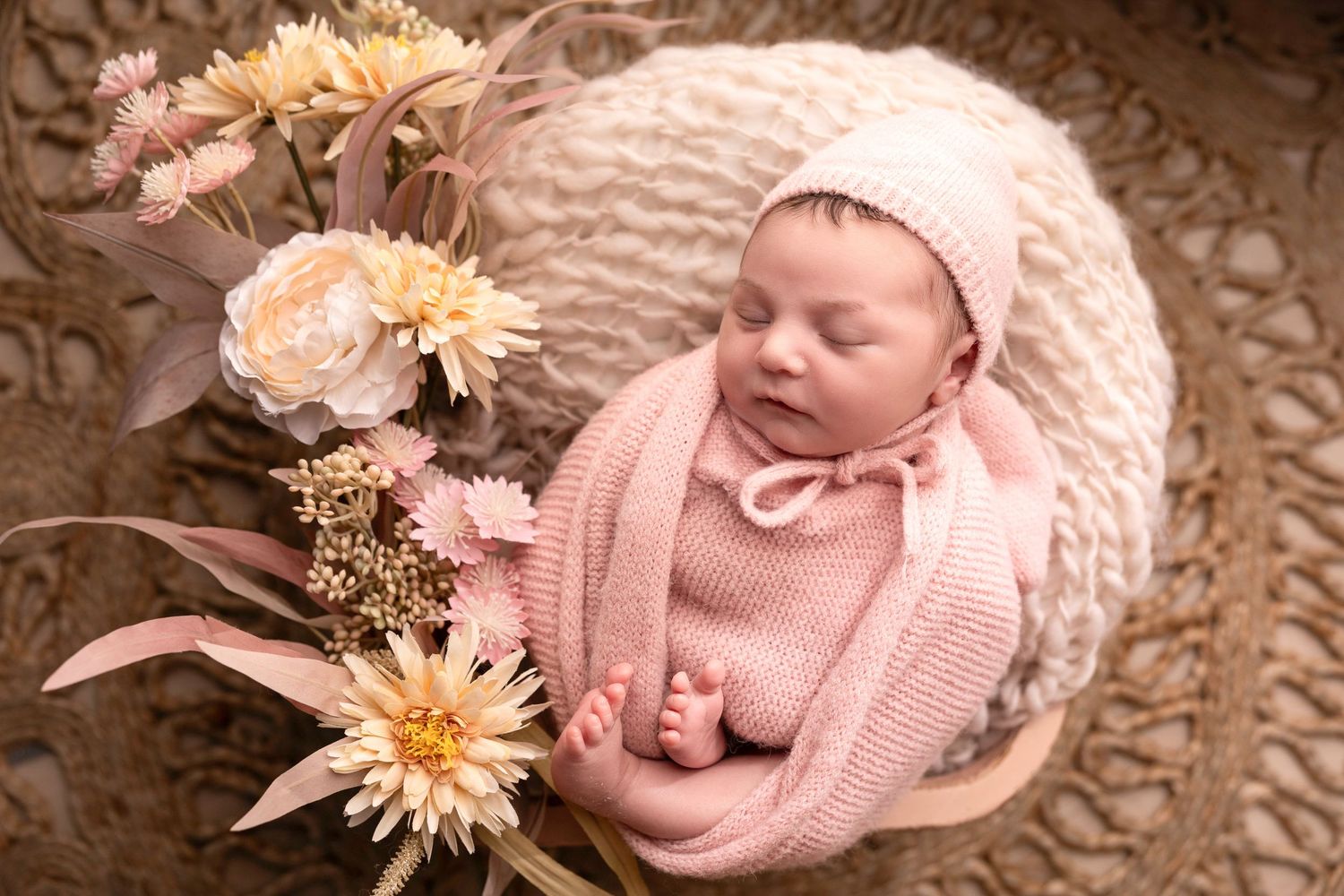 Sleeping newborn in pink wrap surrounded by peachy flowers in basket.