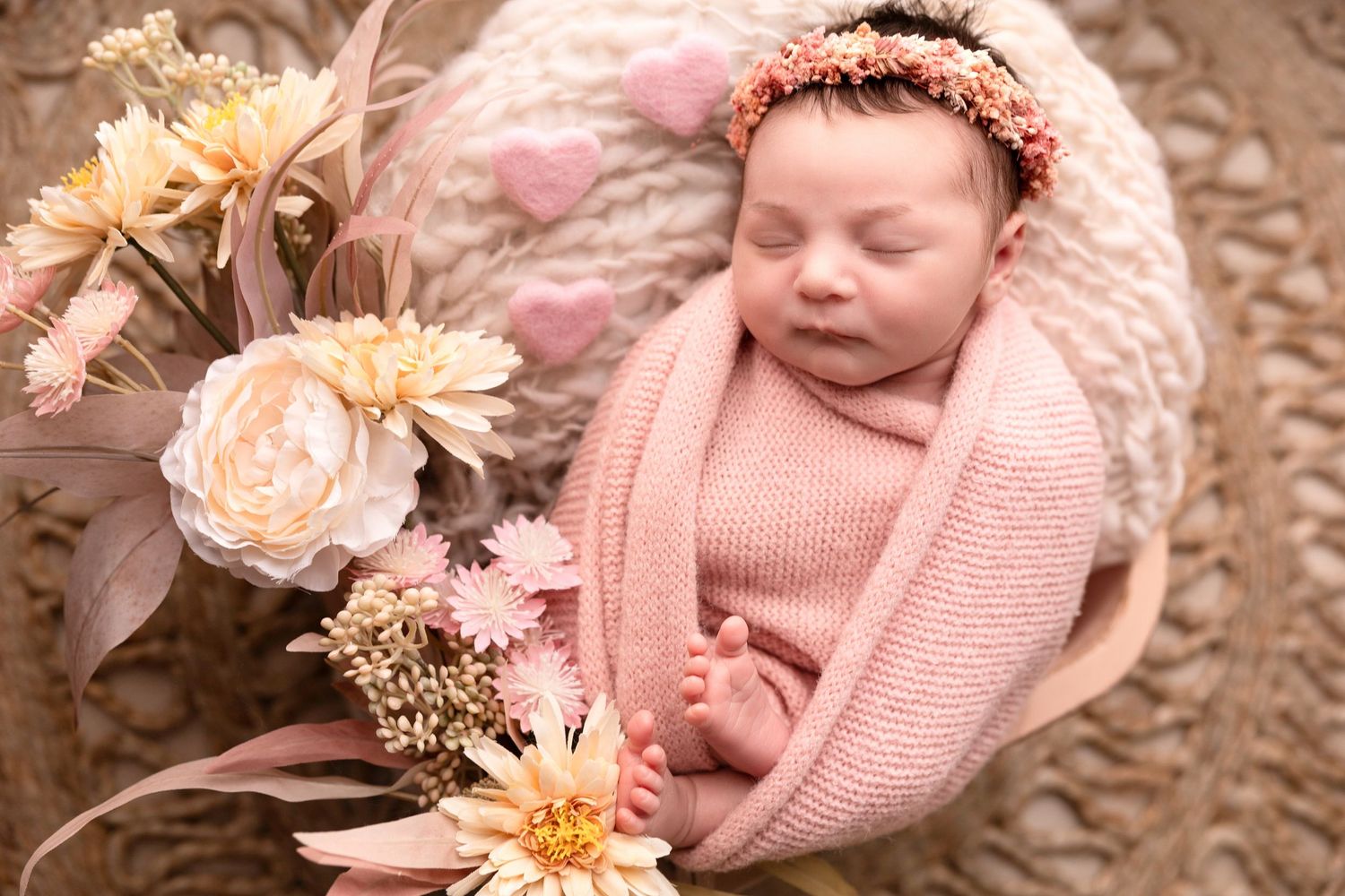 Newborn baby wrapped in pink knit blanket with floral decorations in soft natural lighting.