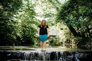 Person relaxes while sitting on top of a small waterfall area surrounded by lush green forest scenery.