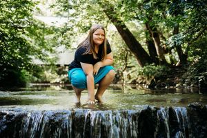 A person in athletic wear crouches beside a small waterfall in a lush forest setting during a summer day.