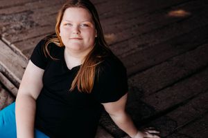 Someone with long auburn hair wearing a black t-shirt sits on a wooden floor in an indoor portrait setting.